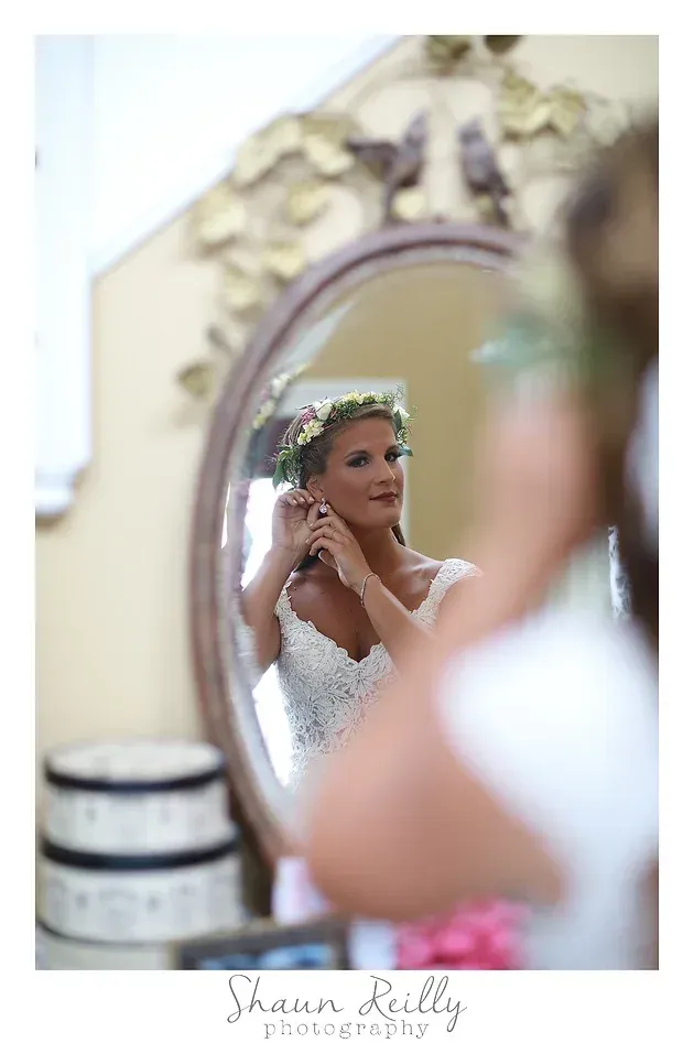 Bride adjusting earring in mirror, wearing a white lace dress and floral hairpiece.