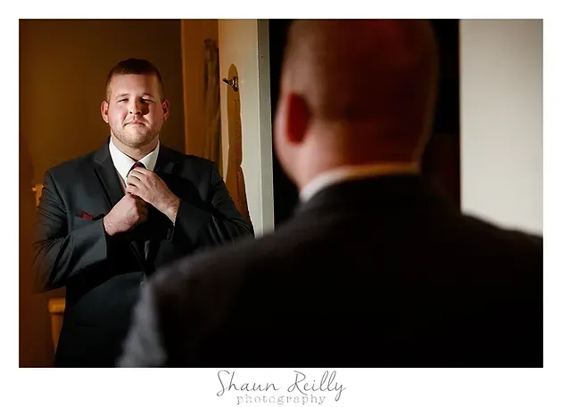Man in dark suit adjusting tie while looking in mirror in a warmly lit room