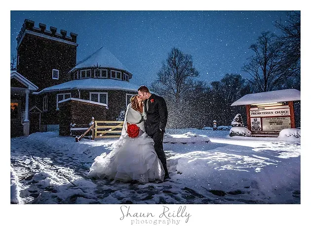 Bride and groom embracing in the snow at night, with lit house and gazebo in the background