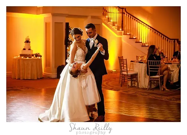 Bride and groom dancing in a warmly lit ballroom with a decorated wedding cake nearby