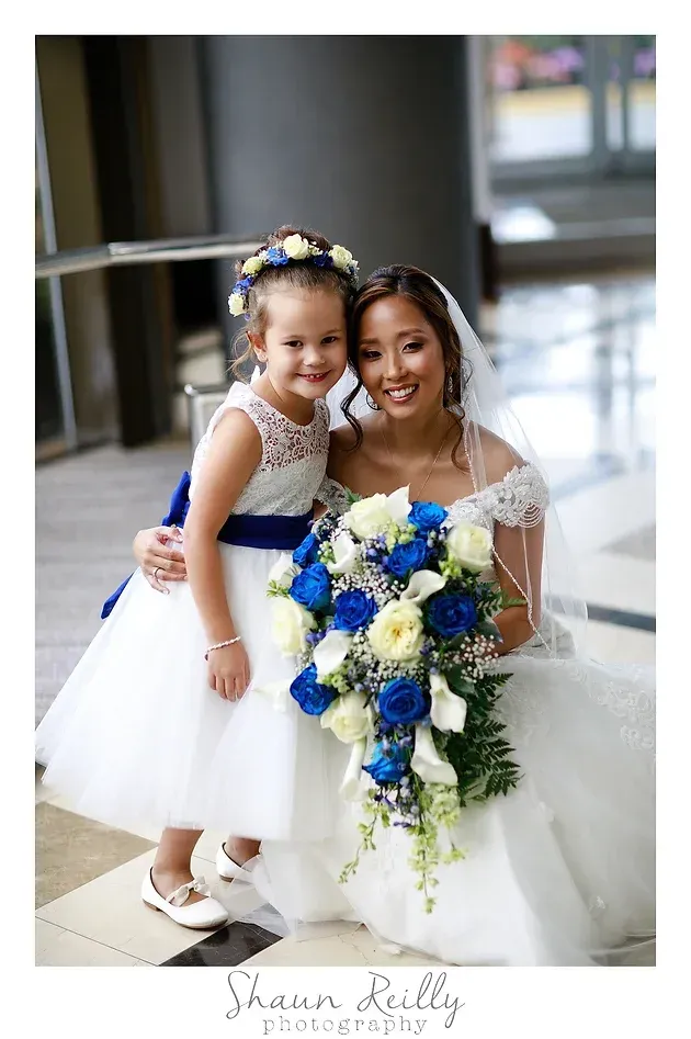 Bride kneels beside a young girl holding a blue-and-white bouquet in a bright indoor hallway