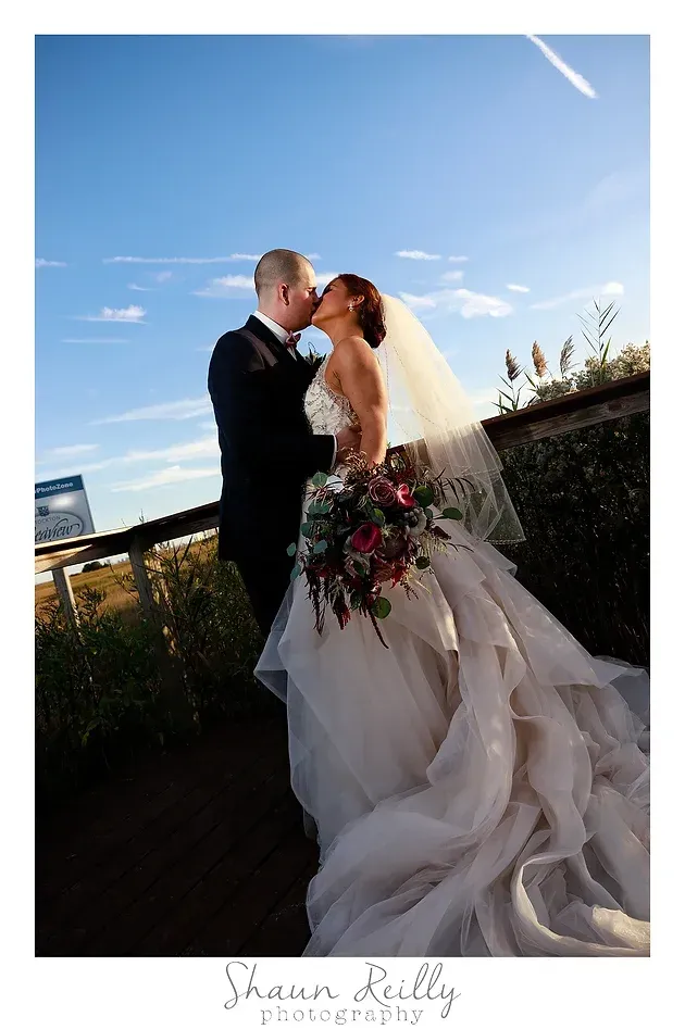 Bride and groom kissing outdoors under a blue sky, bride in a flowing white gown holding a bouquet