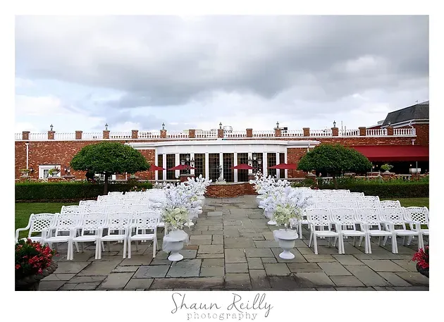 Outdoor wedding ceremony setup with rows of white chairs facing a red-brick building under cloudy skies