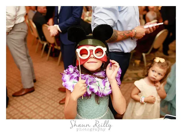 Child in Mickey Mouse ears and red glasses posing with a purple lei at a party, with another child in the background