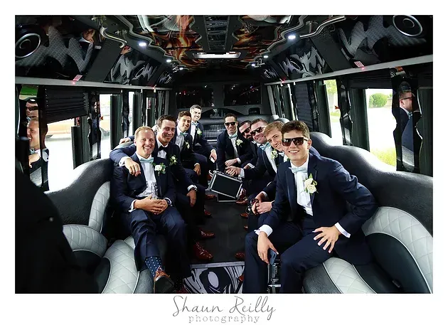 Group of people in formal attire seated inside a black-and-white party bus, smiling and holding drinks.