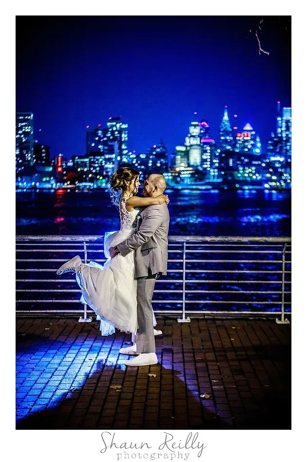 Couple embracing on a waterfront promenade at night, with city skyline lights in the background.