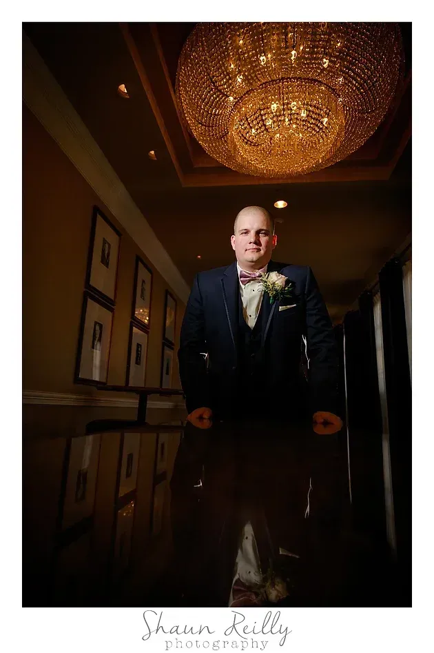 Man in a dark suit standing in a warmly lit hallway beneath a large chandelier