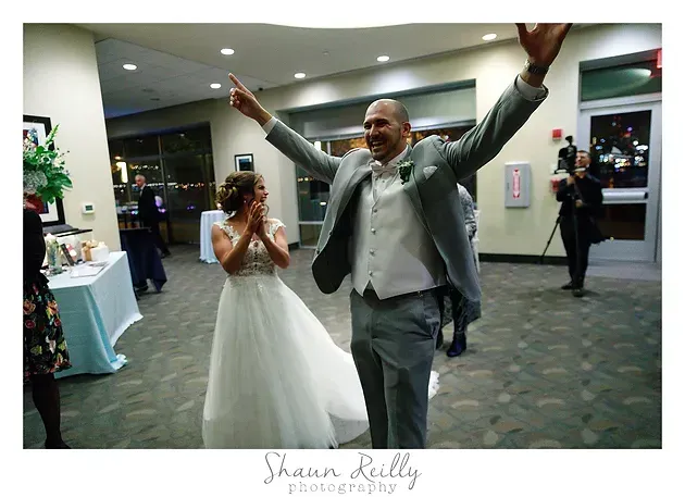 Wedding guest in gray suit celebrates with arms raised as bride in white dress stands nearby indoors