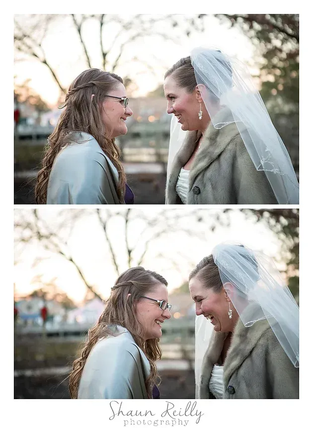Two women in white wedding veils smile at each other outdoors in soft sunlight.