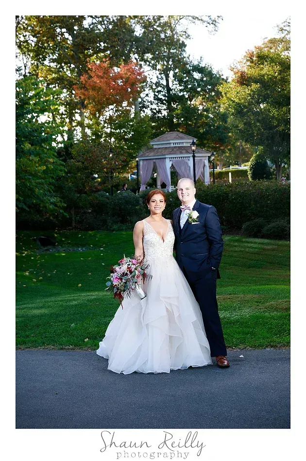 Bride and groom standing together in a garden, with a gazebo and autumn trees behind them.