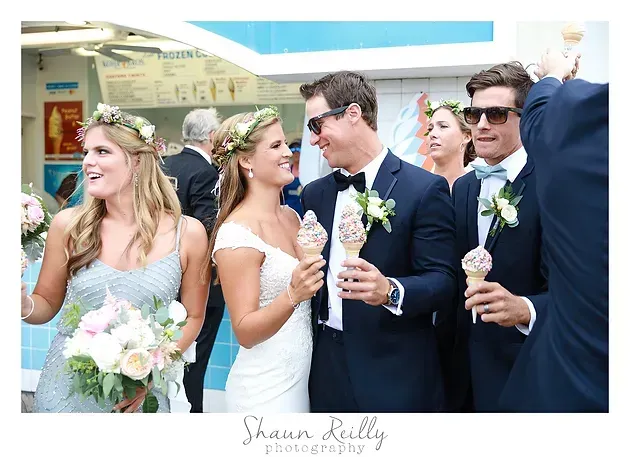 Wedding party in navy suits and white dresses holding roses, smiling outdoors in front of a plane.