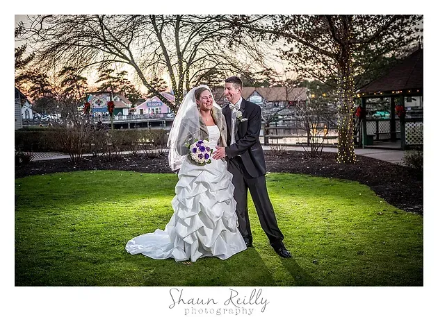 Bride and groom posing outdoors on a lawn at dusk, with trees and buildings in the background.