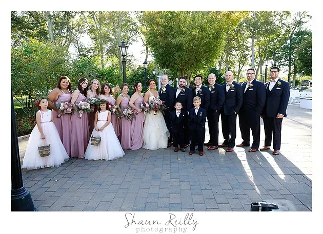 Wedding party posing outdoors in pink and black formal attire, holding bouquets and a flower girl in front.