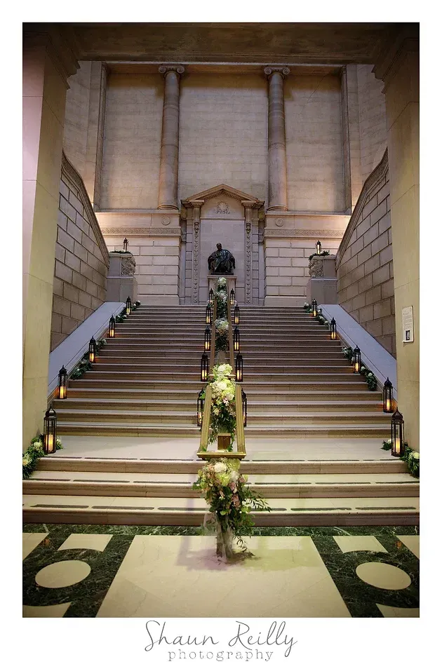 Grand staircase with floral arrangements leading to a statue in a stone hall
