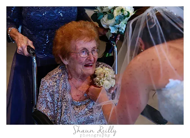 Bride in veil smiling at elderly woman holding flowers during a wedding ceremony