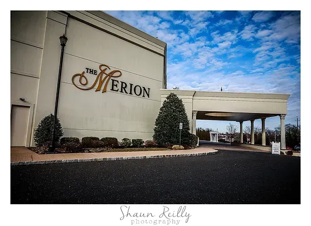 The Merion building entrance with white facade, evergreen shrubs, and a blue sky overhead