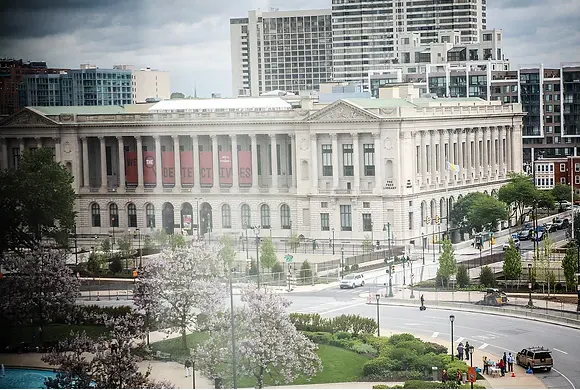 Large white neoclassical building beside a busy road, with city high-rises and trees in the background.