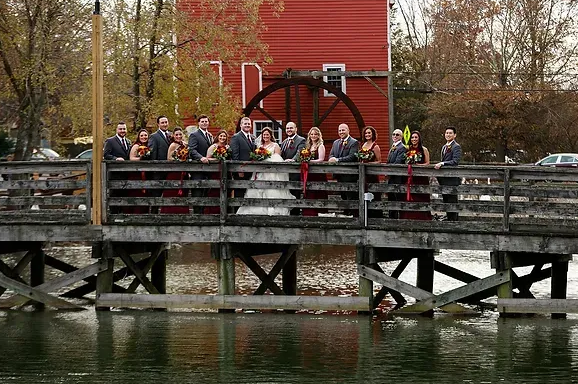 People on a wooden bridge by a red building, gathered near a large black water wheel over a river.