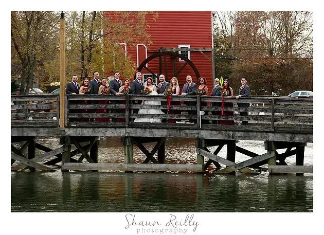 Group of people standing on a wooden bridge beside a red mill over calm water