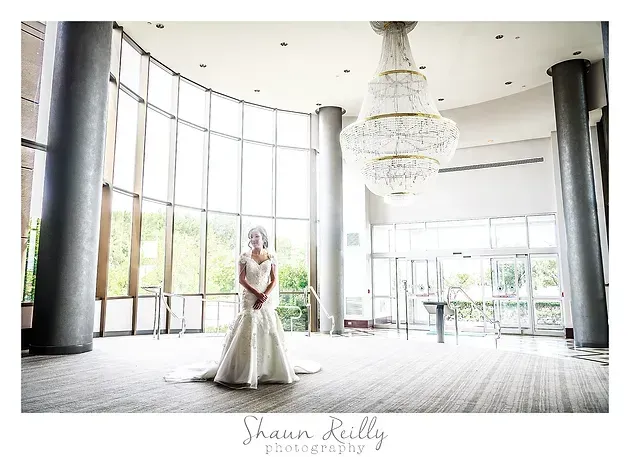 Bride in a white gown standing in a bright modern lobby with large windows and a chandelier