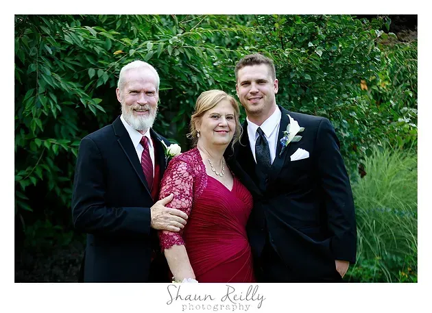 Three people posing outdoors in formal attire, smiling in front of green foliage.
