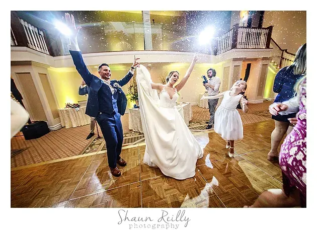 Couple dancing at a wedding reception, arms raised on a polished wooden floor under warm lights