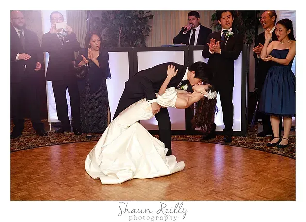 Bride dips groom in a dance at a wedding reception, guests applauding nearby.