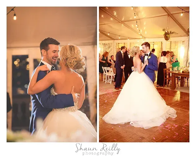 Wedding couple dancing under warm string lights in a reception tent, bride in white gown and groom in blue suit.