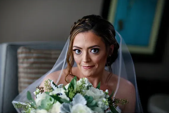 Bride holding a white bouquet, wearing a veil and smiling indoors.
