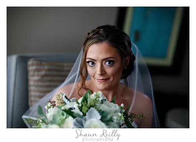 Bride holding a white and green bouquet, wearing a veil and smiling at the camera indoors
