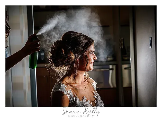 Woman getting hair sprayed backstage, lit warmly in profile.