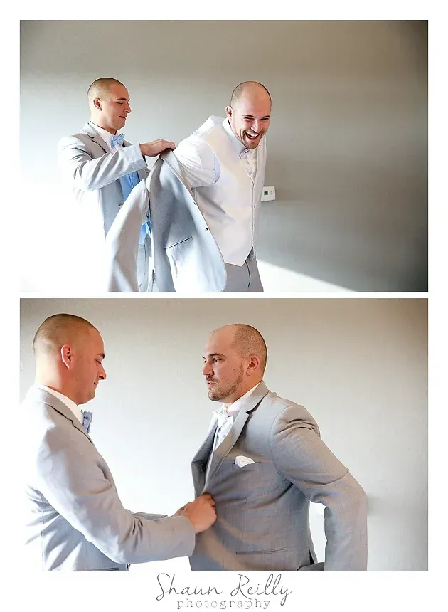 Two men in light suits helping one adjust his jacket in a bright room.