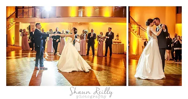 Bride and groom dance in a warmly lit wedding reception hall as guests watch and applaud.