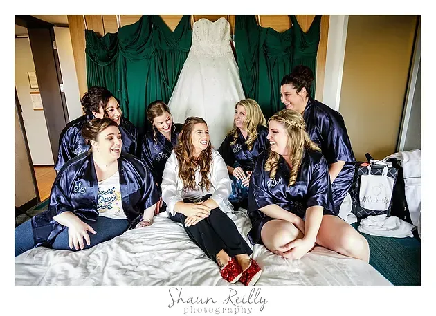 Bridesmaids in navy robes sit on a bed around a white wedding dress hanging behind them.
