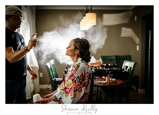 Stylist spraying hairspray on seated woman in floral robe during a salon prep session