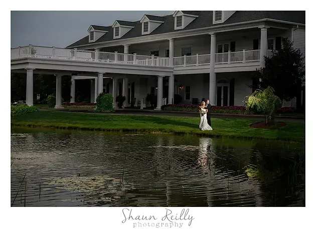 Bride in white on a lawn by a large gray mansion beside a pond