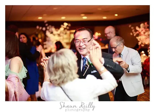 Guests dancing at a warm-lit indoor event, with a smiling man in a tie at center.