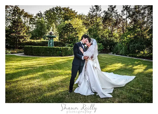 Bride and groom embracing in a garden, with a fountain and trees in the background.