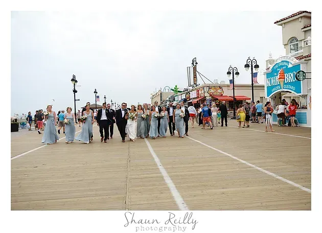 Wedding party posing on a wooden boardwalk by the ocean, with guests and buildings in the background