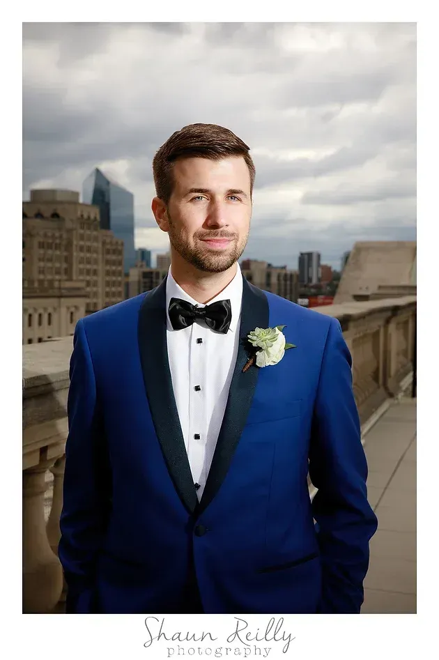 Man in a royal blue tuxedo posing on a rooftop with a cloudy city skyline behind him