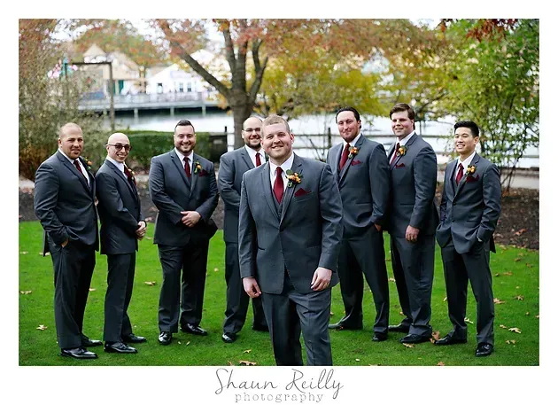 Seven people in dark suits posing outdoors on a lawn with autumn trees and a river backdrop.