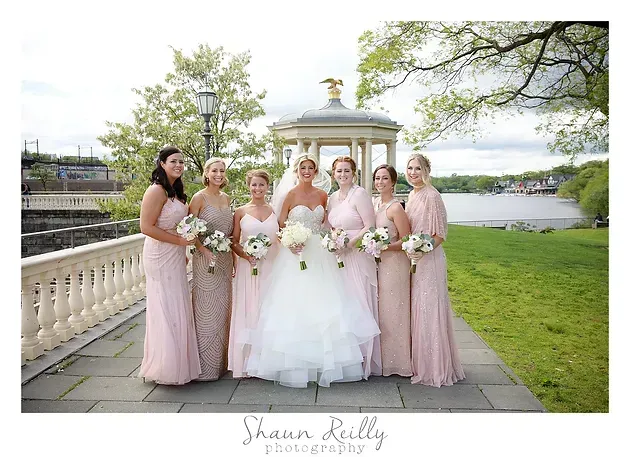 Bridesmaids and bride in pale pink and white dresses pose on a lakeside lawn by a gazebo