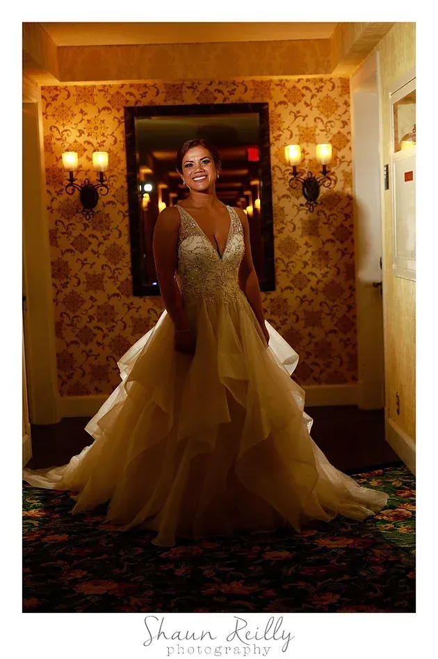 Woman in a gold gown standing in an ornate hallway, smiling under warm lights.