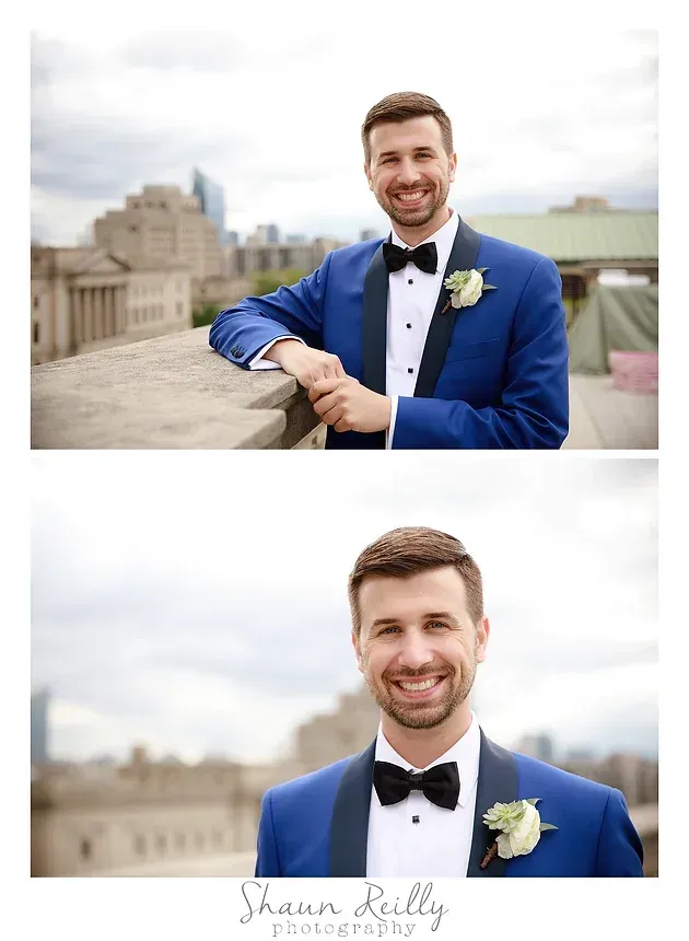 Smiling groom in blue tuxedo with black bow tie, leaning on a ledge outdoors with city buildings behind him