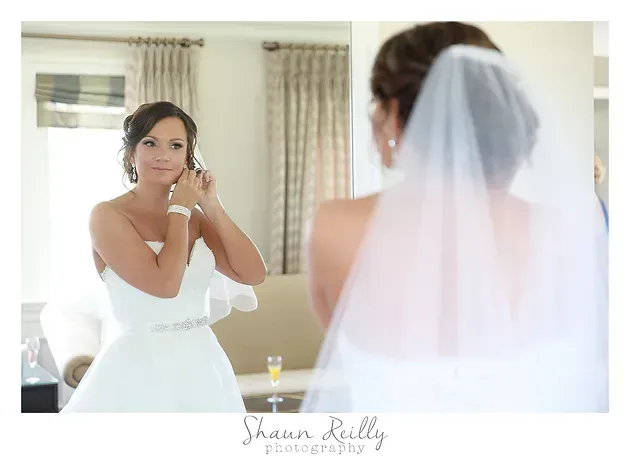 Bride in white dress and veil adjusting earrings in a mirror before the ceremony