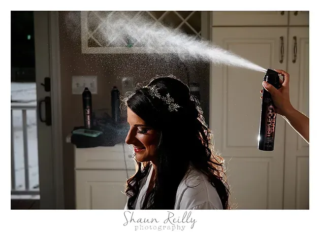 Woman smiling as hairspray is applied in a bathroom, with mist spraying across the mirror.