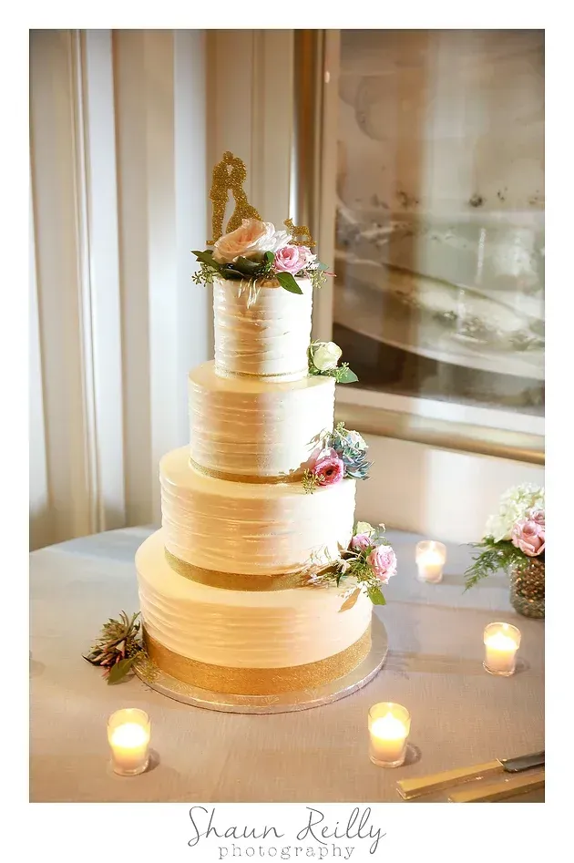 Four-tier ivory wedding cake with gold accents and pink flowers, surrounded by candles in a bright room