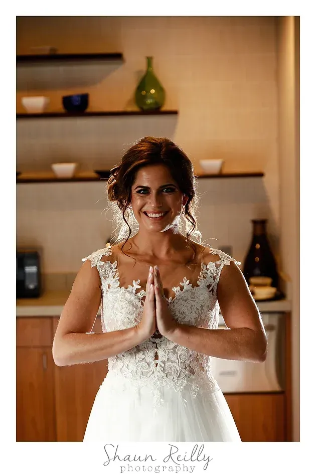 Woman in a white lace dress smiling with hands in prayer pose indoors near shelves