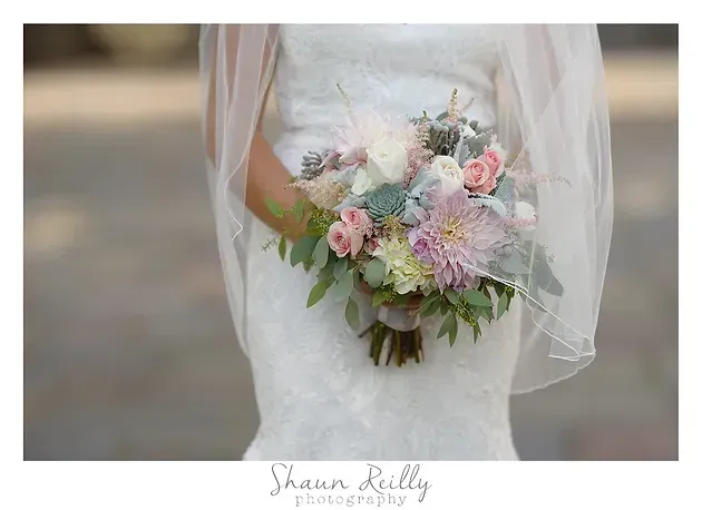 Bridal bouquet of blush and white flowers with greenery, held beneath a sheer veil
