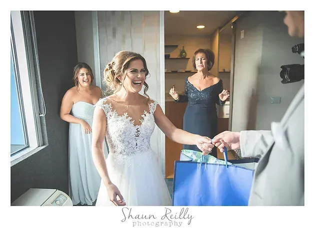 Bride in a white lace dress smiling while someone hands her a blue gift bag, with two women behind her.